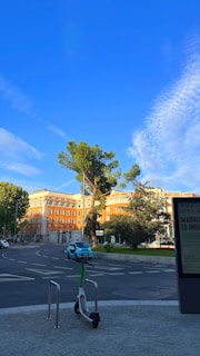 A tranquil urban scene with a bright blue sky and thin white clouds. In the foreground, there is a parked electric scooter on a paved sidewalk. The middle ground features a blue compact car driving on a roundabout. Surrounding the road, well-maintained greenery and large trees can be seen. The background is dominated by a large brick building with numerous windows, standing under the clear sky.