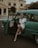 A woman dressed in retro fashion sits in the open door of an antique green car, wearing a headscarf, sunglasses, and a white dress paired with pearls. The background features a classic building with architectural details, conveying a vintage aesthetic.