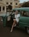 A woman dressed in retro fashion sits in the open door of an antique green car, wearing a headscarf, sunglasses, and a white dress paired with pearls. The background features a classic building with architectural details, conveying a vintage aesthetic.