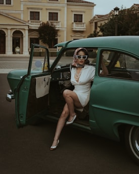 A woman dressed in retro fashion sits in the open door of an antique green car, wearing a headscarf, sunglasses, and a white dress paired with pearls. The background features a classic building with architectural details, conveying a vintage aesthetic.