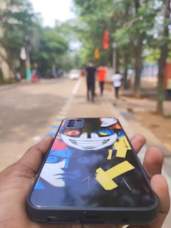 A hand holds a smartphone with a case depicting vibrant, stylized artwork of an animated character. The foreground focuses on the phone, showing a colorful design with expressive eyes and bold lines. In the background, a blurred view of a tree-lined street with people walking and colorful flags can be seen.