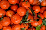 A close-up of a pile of bright orange tangerines, some with green leaves still attached, creating a fresh and vibrant display.