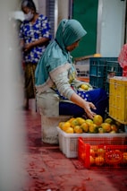 A woman wearing a headscarf and patterned clothes is seated on a low stool, sorting or arranging a pile of oranges in plastic crates. The floor is red-tiled and there are crates filled with oranges scattered around. Another person in the background is wearing a mask and a blue patterned shirt.