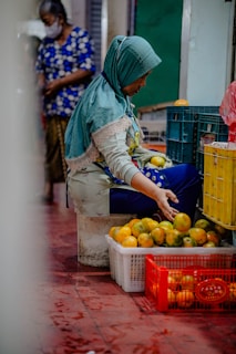 A woman wearing a headscarf and patterned clothes is seated on a low stool, sorting or arranging a pile of oranges in plastic crates. The floor is red-tiled and there are crates filled with oranges scattered around. Another person in the background is wearing a mask and a blue patterned shirt.