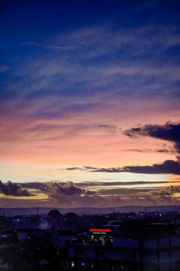 A vibrant sunset with a gradient from deep blue at the top to pink and orange near the horizon, clouds scattered across the sky. Below, a silhouetted cityscape with recognizable buildings and lights, including a clearly visible sign of a hotel.