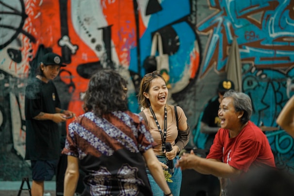 A group of people are interacting joyfully in front of a colorful graffiti-covered wall. One woman in the center is laughing and holding a microphone. The scene conveys a lively and energetic atmosphere.