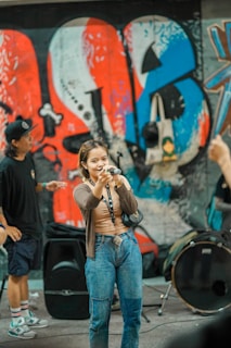 A young woman holds a microphone and appears to be speaking or singing in front of a vibrant graffiti wall. She is casually dressed in jeans and a brown top. In the background, musical equipment, including a drum, is visible, and another person wearing a cap stands nearby.