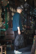 A young person wearing a blue and green checkered shirt and a gray cap stands in a shop filled with various items. They hold a black backpack in their hand. The shop is dimly lit, and numerous decorative pieces are displayed in the background.