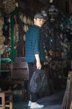 A young person wearing a blue and green checkered shirt and a gray cap stands in a shop filled with various items. They hold a black backpack in their hand. The shop is dimly lit, and numerous decorative pieces are displayed in the background.