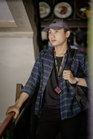 A person is standing on a staircase, wearing a blue and green plaid shirt with a cap, holding onto the railing. They have a lanyard and a small red device around their neck, and a backpack strap is visible on their shoulder. Behind them, a wall with some vintage-style round signs in the background creates a slightly rustic atmosphere.