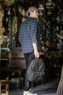 A person is standing in an antique shop or similar setting, wearing a blue and green plaid shirt, black pants, white sneakers, and a gray cap while holding a textured gray backpack. The surroundings include a variety of hanging objects, such as masks and lamps, and there is a wooden chair nearby.