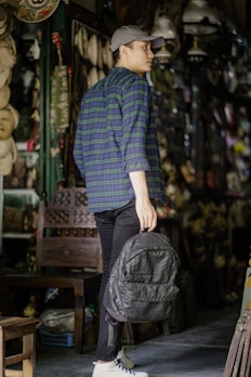 A person is standing in an antique shop or similar setting, wearing a blue and green plaid shirt, black pants, white sneakers, and a gray cap while holding a textured gray backpack. The surroundings include a variety of hanging objects, such as masks and lamps, and there is a wooden chair nearby.