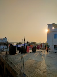 A rooftop terrace with laundry hanging on clotheslines, set against the backdrop of a sunset. The sunlight casts long shadows across the cement floor. In the background, several buildings are visible, and the distant landscape suggests a body of water.