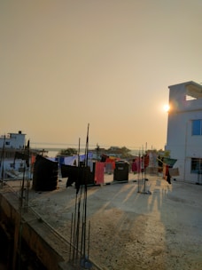 A rooftop terrace with laundry hanging on clotheslines, set against the backdrop of a sunset. The sunlight casts long shadows across the cement floor. In the background, several buildings are visible, and the distant landscape suggests a body of water.