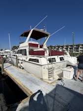 A white motorboat with maroon accents is docked at a marina. The boat is named 'Castle X' and has a ladder leading to an upper deck. It is tied to the dock with several ropes, and a person in shorts is visible standing nearby. In the background, there are more boats and waterfront buildings under a clear blue sky.