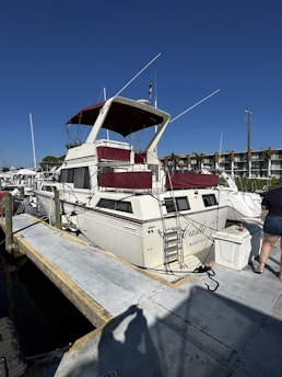 A white motorboat with maroon accents is docked at a marina. The boat is named 'Castle X' and has a ladder leading to an upper deck. It is tied to the dock with several ropes, and a person in shorts is visible standing nearby. In the background, there are more boats and waterfront buildings under a clear blue sky.