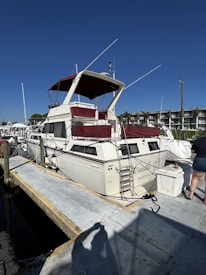 A white motorboat with maroon accents is docked at a marina. The boat is named 'Castle X' and has a ladder leading to an upper deck. It is tied to the dock with several ropes, and a person in shorts is visible standing nearby. In the background, there are more boats and waterfront buildings under a clear blue sky.