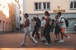 A group of young people walks energetically along a street. One person is sitting in a shopping cart, and the others are walking alongside, some carrying bags. The building behind them features the signage 'HUBER'. The street is lined with buildings, and a parked car is partially visible.