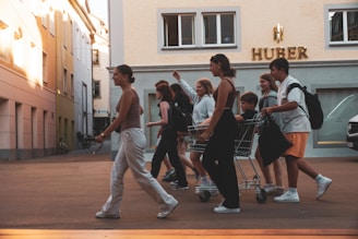 A group of young people walks energetically along a street. One person is sitting in a shopping cart, and the others are walking alongside, some carrying bags. The building behind them features the signage 'HUBER'. The street is lined with buildings, and a parked car is partially visible.