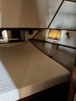 A cozy cabin interior features a neatly made bed with white linens set in a small, enclosed space. Wooden trim and a small window with a striped curtain allow warm light to filter in. A wall-mounted lamp provides additional lighting nearby.