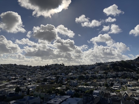 A neighborhood sprawls across rolling hills under a sky filled with large, fluffy clouds. The sky is bright, indicating a sunny atmosphere. Houses of various sizes and styles cover the landscape, with streets weaving through them, creating an urban tapestry.
