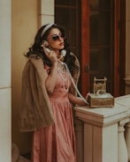 A woman in a vintage setting is wearing a fur stole and a pink satin dress, accessorized with pearls and sunglasses. She is holding a golden retro telephone to her ear, standing by a stone balustrade against a backdrop of wooden doors.