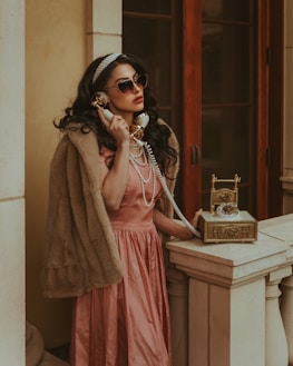 A woman in a vintage setting is wearing a fur stole and a pink satin dress, accessorized with pearls and sunglasses. She is holding a golden retro telephone to her ear, standing by a stone balustrade against a backdrop of wooden doors.