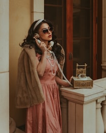 A woman in a vintage setting is wearing a fur stole and a pink satin dress, accessorized with pearls and sunglasses. She is holding a golden retro telephone to her ear, standing by a stone balustrade against a backdrop of wooden doors.