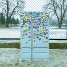 A utility box covered in various colorful stickers is positioned in front of a chain-link fence. The ground is paved, and the background shows bare trees and a partly snowy landscape with overcast weather.