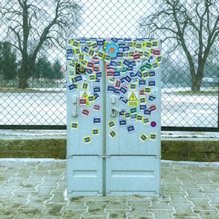 A utility box covered in various colorful stickers is positioned in front of a chain-link fence. The ground is paved, and the background shows bare trees and a partly snowy landscape with overcast weather.