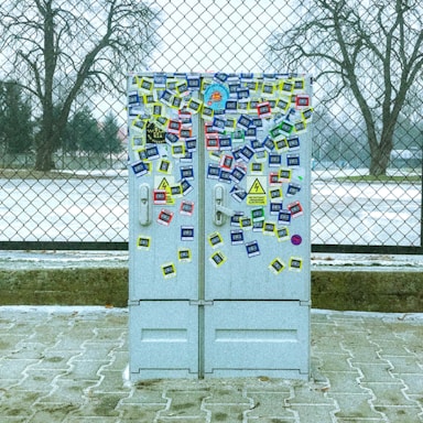 A utility box covered in various colorful stickers is positioned in front of a chain-link fence. The ground is paved, and the background shows bare trees and a partly snowy landscape with overcast weather.