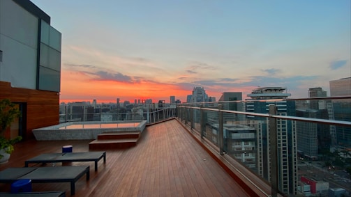 A rooftop terrace with wooden flooring overlooks a cityscape during sunset. The sky displays vibrant hues of orange and pink, contrasting with the silhouettes of modern skyscrapers. A glass railing runs along the edge, and there are lounge chairs with a small table near a jacuzzi in the corner.