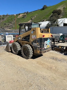 A construction vehicle, specifically a skid steer loader, is parked on a dirt surface. The vehicle is primarily yellow with visible dirt and scuff marks, indicating frequent use. Surrounding the loader are various industrial objects, including a fence, containers, and a hill covered in green vegetation in the background.