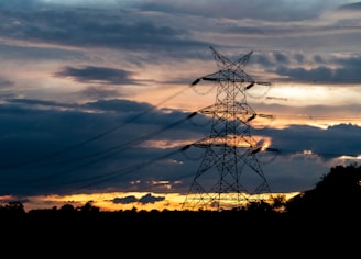 A large electricity pylon stands silhouetted against a dramatic sky filled with dark clouds and an orange-hued sunset.