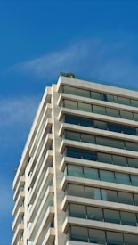 A modern high-rise building with multiple floors and a glass facade is depicted against a clear blue sky. The structure features balconies with glass railings and appears to have some greenery on the rooftop.
