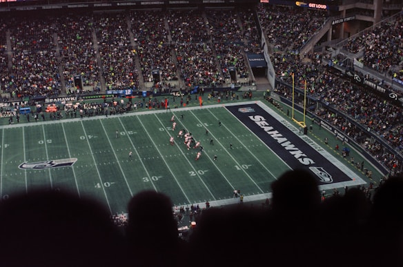 A crowded football stadium with fans filling the stands, a grass field with visible yard markers, and players actively engaged in a game near the goal line. The end zone is marked with 'Seahawks' and team logos are present on the field.