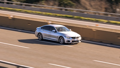 A silver sedan is driving along a highway, with motion blur indicating speed. The car is in the left lane, and the background shows a concrete barrier, grass, and trees.