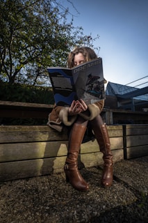 A person sitting on a wooden bench, engrossed in reading a magazine titled 'Dreams'. The scene is set outdoors with trees and a building in the background. The person is wearing a coat and knee-high brown boots.