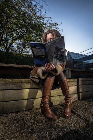 A person sitting on a wooden bench, engrossed in reading a magazine titled 'Dreams'. The scene is set outdoors with trees and a building in the background. The person is wearing a coat and knee-high brown boots.