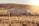 A large semi-truck with a white trailer is traveling on a deserted road through a dry, grassy landscape with rolling hills in the background. The sun is setting or rising, casting a warm glow over the scene.