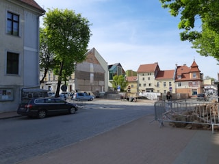 A street in a small town with buildings featuring traditional and modern architecture. Cars are parked along the road, and a few people can be seen walking or sitting. Metal barriers are set up, possibly indicating construction or road work. Green trees add a touch of nature to the urban setting.