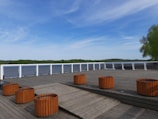 A wooden deck area with cylindrical orange planters placed symmetrically. In the background, there are green trees and a body of water, all under a partly cloudy blue sky.