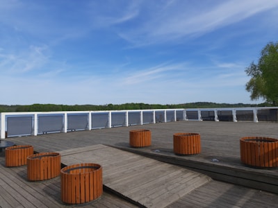 A wooden deck area with cylindrical orange planters placed symmetrically. In the background, there are green trees and a body of water, all under a partly cloudy blue sky.