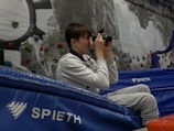A person is sitting on blue gym mats holding a camera to their face, seemingly taking a photograph. They are inside a sports facility with rock climbing holds attached to a wall in the background.