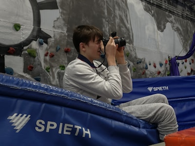 A person is sitting on blue gym mats holding a camera to their face, seemingly taking a photograph. They are inside a sports facility with rock climbing holds attached to a wall in the background.