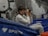 A person is sitting on blue gym mats holding a camera to their face, seemingly taking a photograph. They are inside a sports facility with rock climbing holds attached to a wall in the background.
