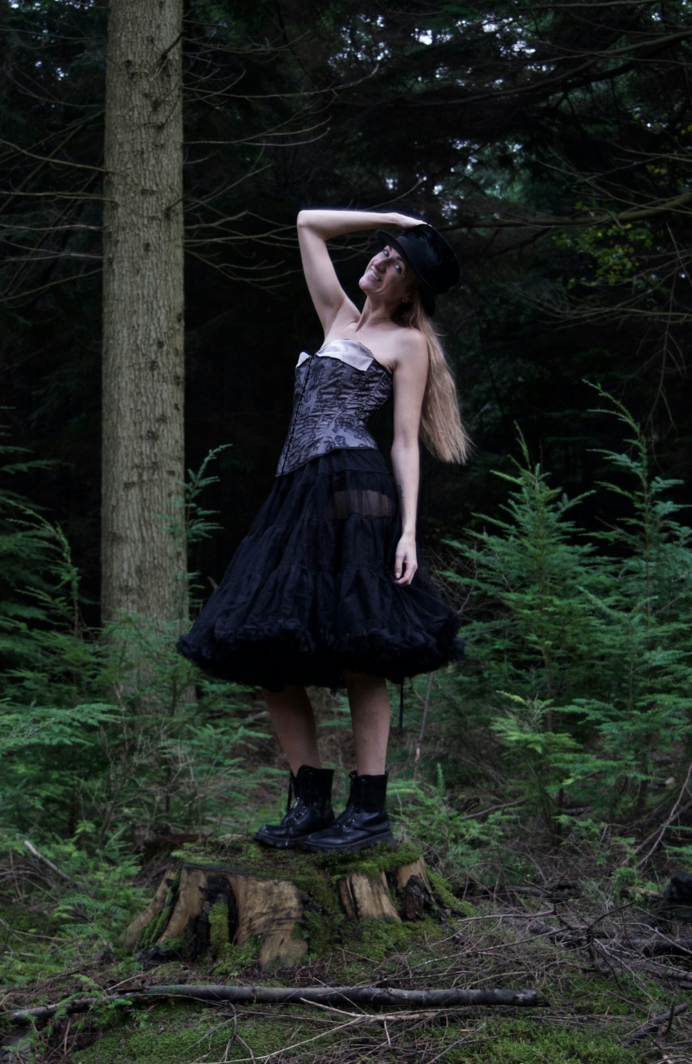 a woman in a black dress and hat standing on a tree stump