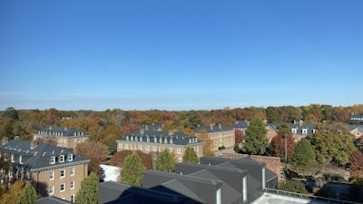 Aerial view of a residential area featuring multiple brick buildings with dark roofs. The surrounding landscape is filled with trees displaying autumn foliage in vibrant shades of orange, red, and green under a clear blue sky.