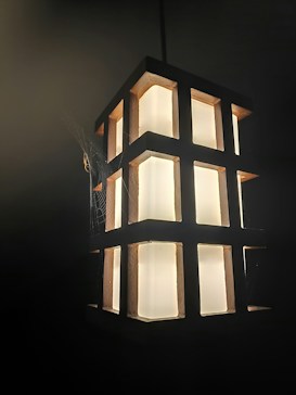 A geometric, rectangular lamp with glowing panels is suspended against a dark background. The lamp is framed by a grid-like structure made of dark material. Several spider webs can be seen clinging to the corners of the lamp, adding a delicate, intricate detail.