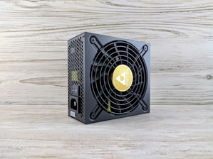 A computer power supply unit with a black casing, featuring a prominent circular fan grille. The center of the fan has a logo with a yellow background. The sides have a honeycomb grill, and there are visible power connectors, including an on/off switch. The unit sits on a light wooden surface with a textured wall in the background.